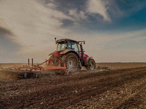 Rode tractor ploegt boerenveld onder bewolkte hemel, land voorbereidend voor magazijnbouwproject. 